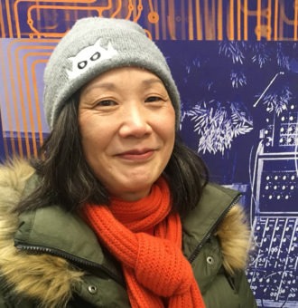 A woman wearing a beanie and orange scarf in front of an orange wall.