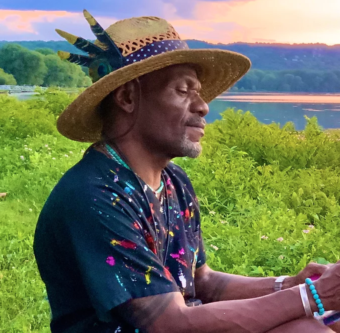 A man in a hat sitting on the grass near a lake.