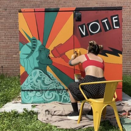 A woman paints a mural on a voting box.