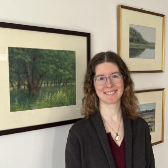 A woman in glasses standing in front of framed paintings.
