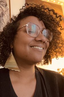 A woman with glasses and curly hair wearing gold earrings.